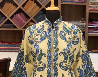 Könnte beinhalten: Eine cremefarbene Tunika mit langen Ärmeln, Stehkragen und einer Knopfleiste auf der Vorderseite. Das Kleidungsstück ist mit einem aufwendigen blauen und grünen Paisley-Stickmuster verziert, das vertikal über Vorderseite und Ärmel verläuft.