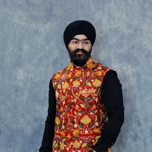 Op de afbeelding: Een man draagt een zwarte turban, een rood vest met bloemenprint, een zwart overhemd met lange mouwen, een zwarte jeans en bruine schoenen.