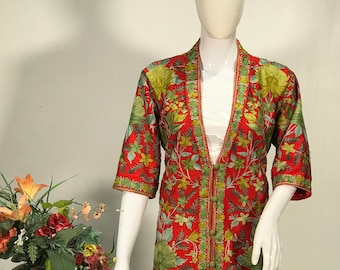 May include: A knee-length red coat featuring an intricate all-over floral embroidery pattern in shades of green, yellow, and orange. The garment has three-quarter length sleeves, a V-neckline, and a front closure secured with four round fabric buttons.