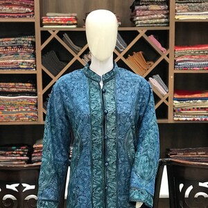 May include: A long, blue embroidered coat displayed on a mannequin. The coat features a detailed floral pattern in shades of blue and green. It has a mandarin collar and button closures down the front. The mannequin stands in front of a wooden shelf filled with textiles.