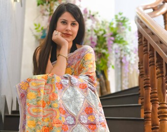 May include: A colorful, embroidered shawl with a floral and paisley design. The shawl features a light gray background with orange, yellow, and purple accents. The intricate embroidery is visible throughout the fabric.