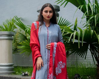 May include: A grey long-sleeved tunic with a mandarin collar and red piping, paired with matching grey trousers. A vibrant red shawl with grey floral embroidery drapes over the shoulders. The text Angad Creations appears at the bottom.