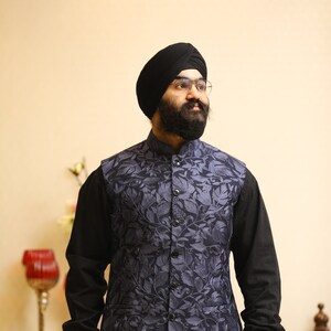Pu&ograve; includere: Un uomo indossa un turbante nero, una camicia nera e un gilet blu con un motivo floreale. Il gilet ha un colletto alla coreana e bottoni sul davanti.