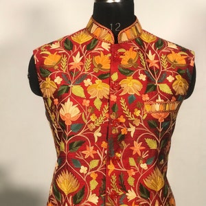 Op de afbeelding: Een rood, mouwloos vest met een bloemenpatroon geborduurd in tinten geel, oranje, groen en wit. Het vest heeft een mandarijnskraag en een knoopsluiting.