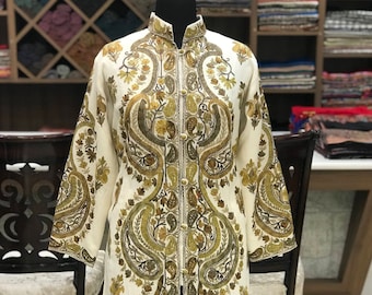 Könnte beinhalten: Ein langärmeliger, cremeweißer Mantel mit einem komplexen, symmetrischen Stickmuster in Olivgrün, Gold und Braun. Das Kleidungsstück verfügt über einen Stehkragen, eine Knopfleiste auf der Vorderseite und eine gerade, knielange Silhouette.
