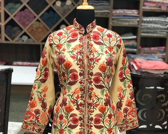 Könnte beinhalten: Eine lange, cremefarbene Jacke mit einem floralen Stickmuster in Rot-, Orange- und Grüntönen. Die Jacke hat einen Stehkragen und einen Reißverschluss vorne.