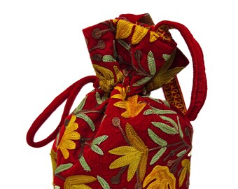 Op de afbeelding: Een rode fluwelen trekkoordtas met een geborduurd bloemenmotief in tinten geel, groen en bruin. De tas heeft een lange, dunne band.