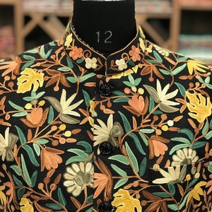 K&ouml;nnte beinhalten: Eine schwarze Weste mit einem floralen Stickmuster in Gr&uuml;n-, Braun- und Gelbt&ouml;nen. Die Weste hat einen Stehkragen und f&uuml;nf Kn&ouml;pfe.