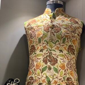 Peut inclure: Un gilet beige sans manches avec un motif floral brod&eacute; dans des tons de brun, vert et jaune. Le gilet a un col mandarin et une fermeture &agrave; boutons. Le gilet est pr&eacute;sent&eacute; sur un mannequin.