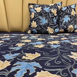 May include: A navy blue bedspread with a pattern of embroidered leaves in shades of blue and beige. Two matching pillowcases with the same leaf pattern are on top of the bedspread.