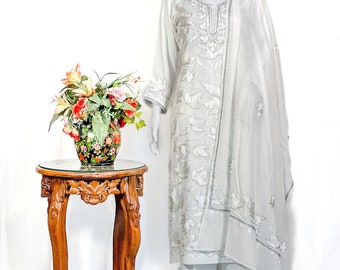 May include: A light gray three-piece outfit consisting of a long-sleeved tunic with silver floral embroidery, matching straight-leg trousers, and a coordinating sheer dupatta draped over the shoulder, all displayed on a mannequin.