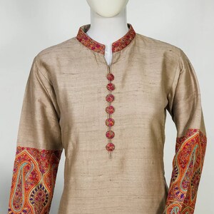 May include: Beige tunic with a mandarin collar and long sleeves. The collar and cuffs feature intricate embroidery in shades of red, orange, blue, and green. The tunic has decorative buttons down the front.