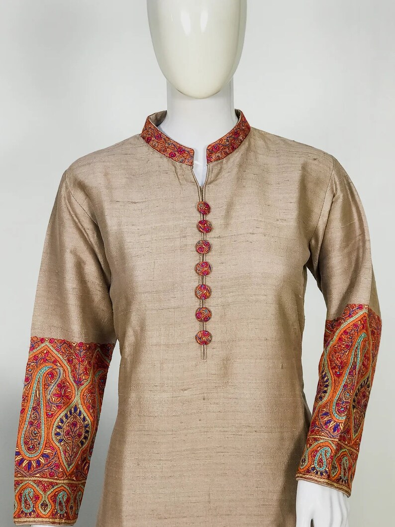 May include: Beige tunic with a mandarin collar and long sleeves. The collar and cuffs feature intricate embroidery in shades of red, orange, blue, and green. The tunic has decorative buttons down the front.