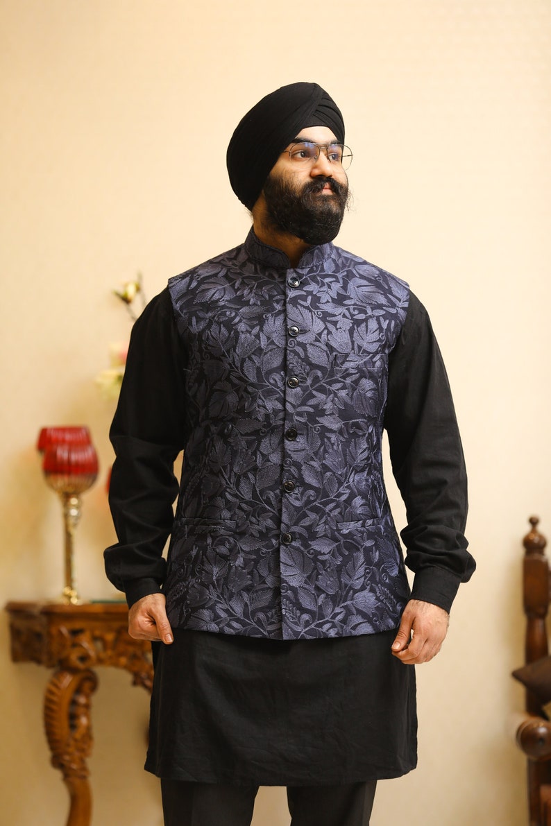 Pu&ograve; includere: Un uomo indossa un turbante nero, una camicia nera e un gilet blu con un motivo floreale. Il gilet ha un colletto alla coreana e bottoni sul davanti.