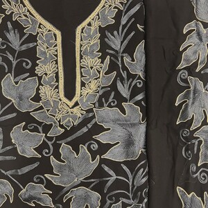 May include: A black tunic featuring intricate embroidery in shades of grey and silver. The design showcases a dense pattern of large leaves and vines covering the fabric, with a prominent gold-toned border outlining the U-shaped neckline.