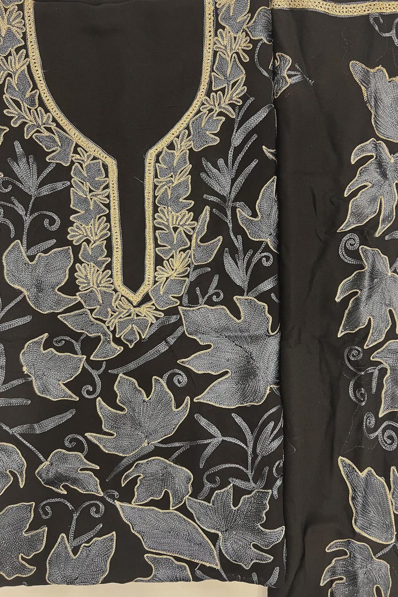 May include: A black tunic featuring intricate embroidery in shades of grey and silver. The design showcases a dense pattern of large leaves and vines covering the fabric, with a prominent gold-toned border outlining the U-shaped neckline.