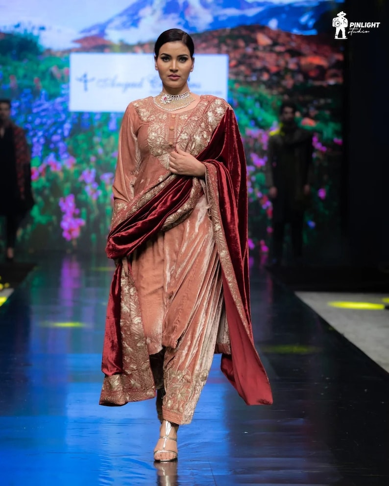 May include: A dusty rose velvet tunic and matching trousers set featuring intricate gold embroidery on the bodice and hem. The outfit is paired with a deep burgundy velvet shawl that has a wide gold embroidered border along the edges.