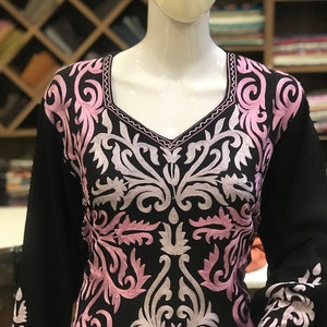 May include: A black long-sleeved tunic featuring an intricate embroidered pattern in shades of light pink and white. The embroidery covers the chest and sleeves, showcasing symmetrical floral and scroll motifs with a square neckline.