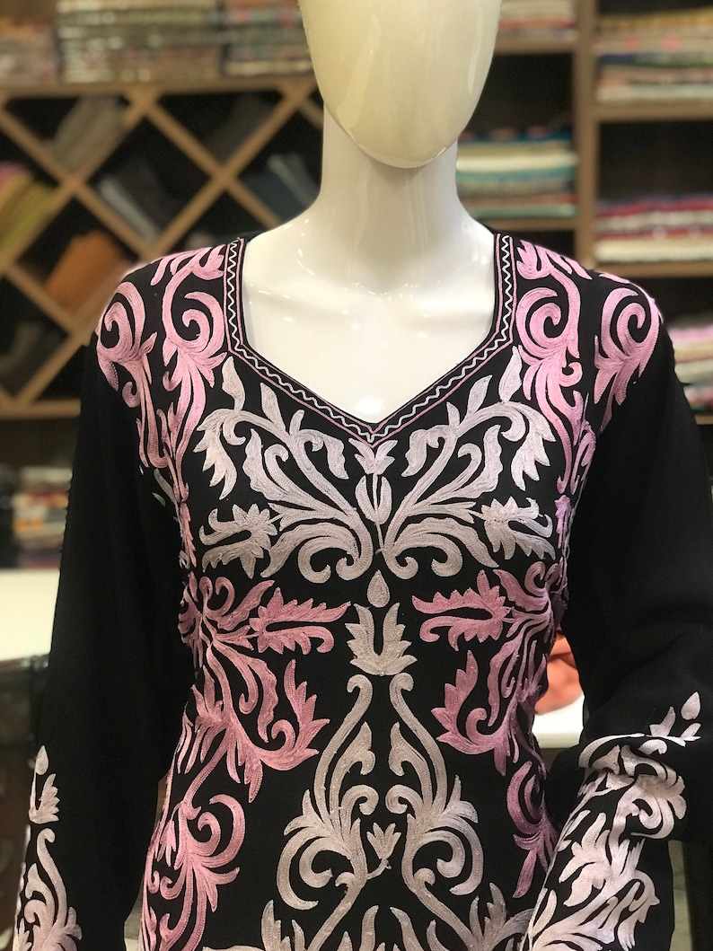 May include: A black long-sleeved tunic featuring an intricate embroidered pattern in shades of light pink and white. The embroidery covers the chest and sleeves, showcasing symmetrical floral and scroll motifs with a square neckline.