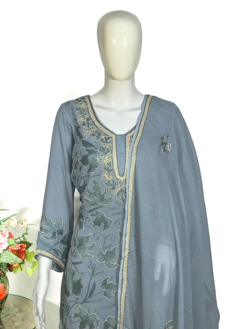 May include: A slate gray long-sleeved tunic featuring intricate dark gray leaf embroidery and gold-toned trim along the neckline and cuffs. The garment is paired with a matching sheer slate gray dupatta draped over one shoulder, showcasing a cohesive design.