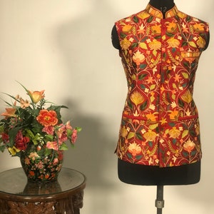 Op de afbeelding: Een rood, mouwloos vest met een bloemenborduurpatroon. Het vest heeft een mandarijnskraag en een knoopsluiting.