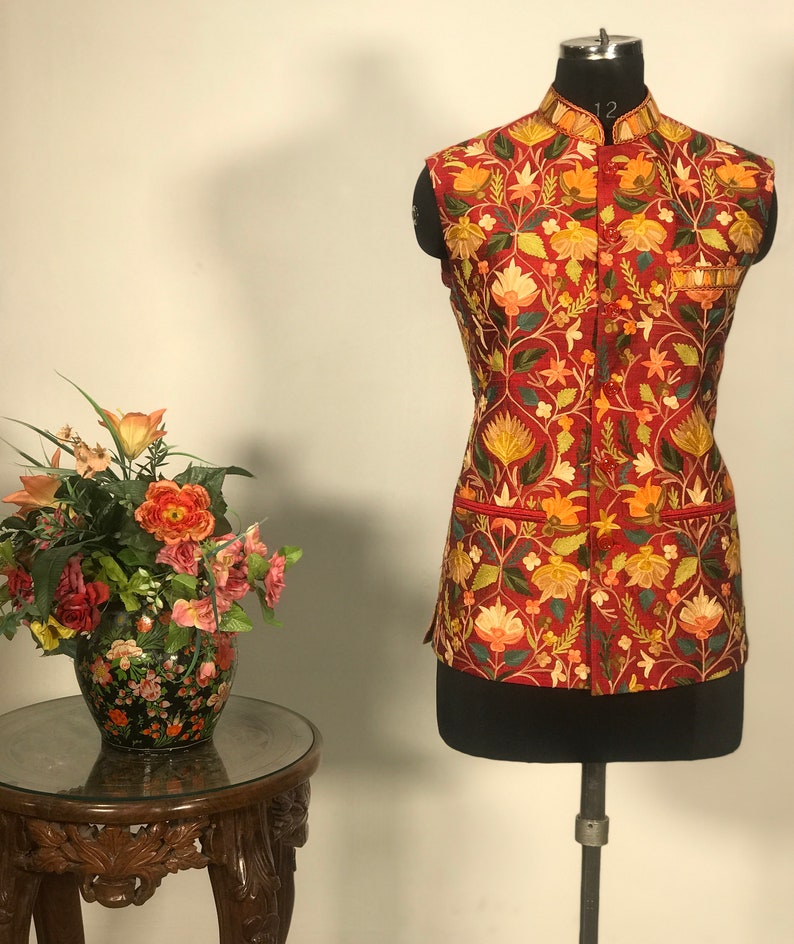 Op de afbeelding: Een rood, mouwloos vest met een bloemenborduurpatroon. Het vest heeft een mandarijnskraag en een knoopsluiting.