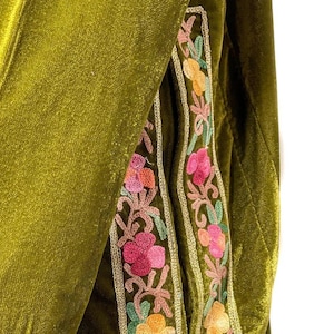 May include: An olive green velvet garment featuring vertical panels of floral embroidery. The embroidery displays pink, orange, and light green thread work, framed by a thin gold-colored metallic border that runs along the edges of the fabric.