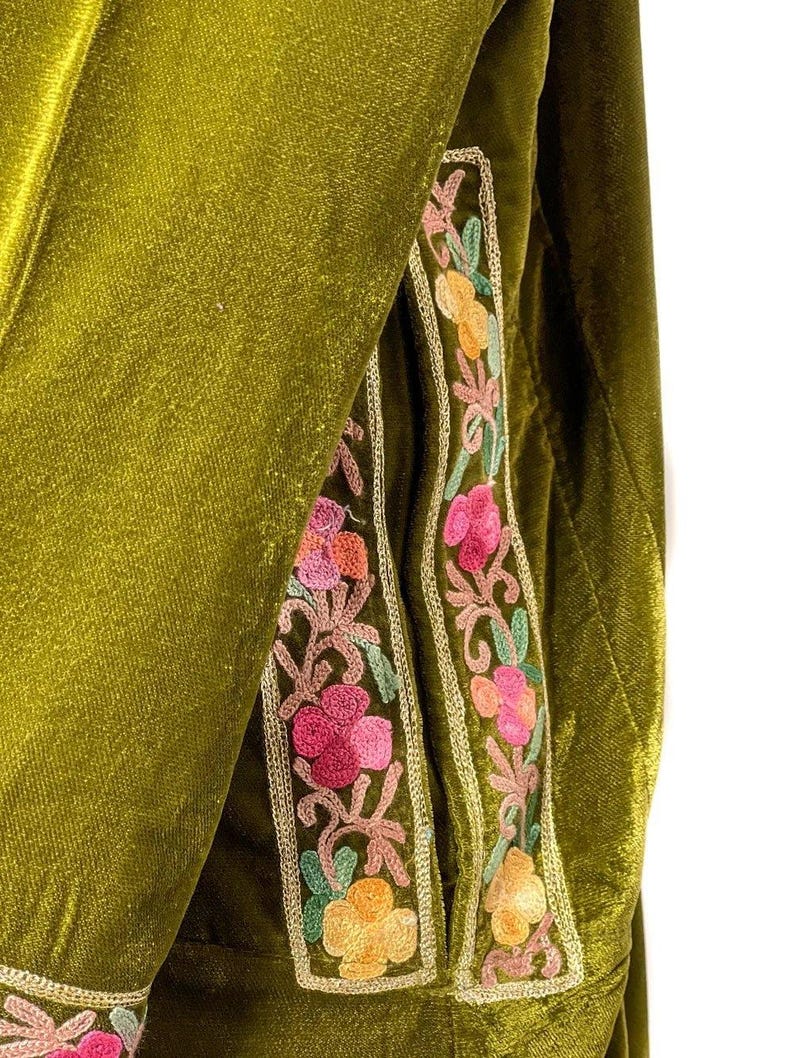 May include: An olive green velvet garment featuring vertical panels of floral embroidery. The embroidery displays pink, orange, and light green thread work, framed by a thin gold-colored metallic border that runs along the edges of the fabric.