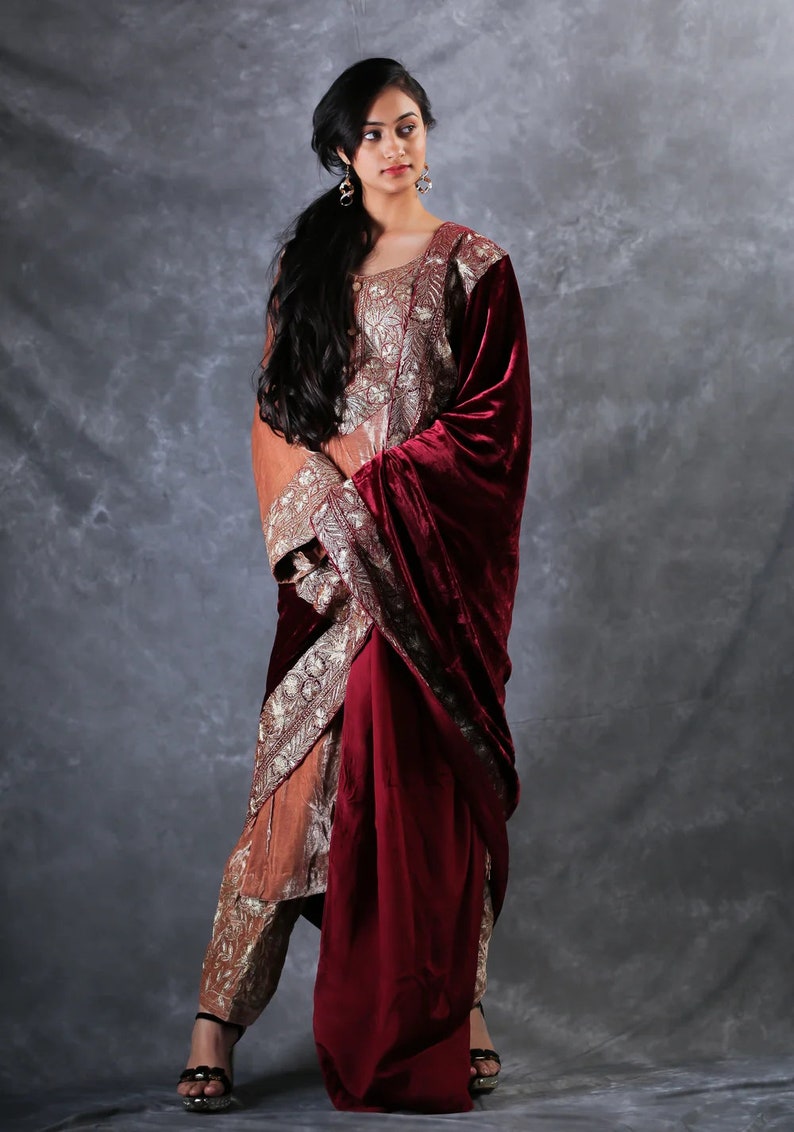 May include: A three-piece ensemble featuring a long-sleeved, copper-colored tunic and matching trousers, both adorned with intricate silver embroidery. A deep burgundy velvet shawl is draped over one shoulder, completing the formal outfit.