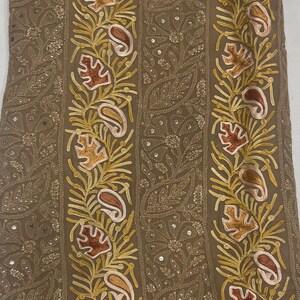 May include: A tan fabric featuring vertical bands of intricate embroidery. The design includes yellow, orange, and pink leaf and paisley motifs interspersed with small, reflective sequins. The background fabric has a subtle, tonal floral pattern.