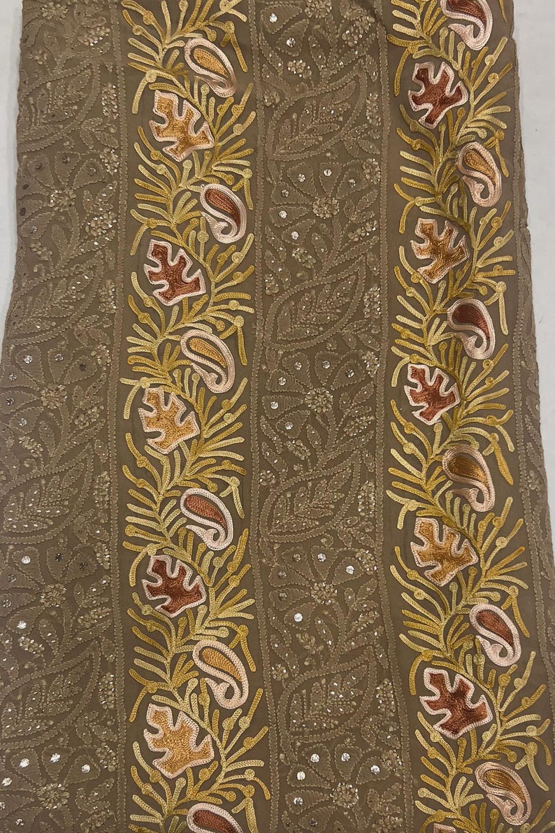 May include: A tan fabric featuring vertical bands of intricate embroidery. The design includes yellow, orange, and pink leaf and paisley motifs interspersed with small, reflective sequins. The background fabric has a subtle, tonal floral pattern.