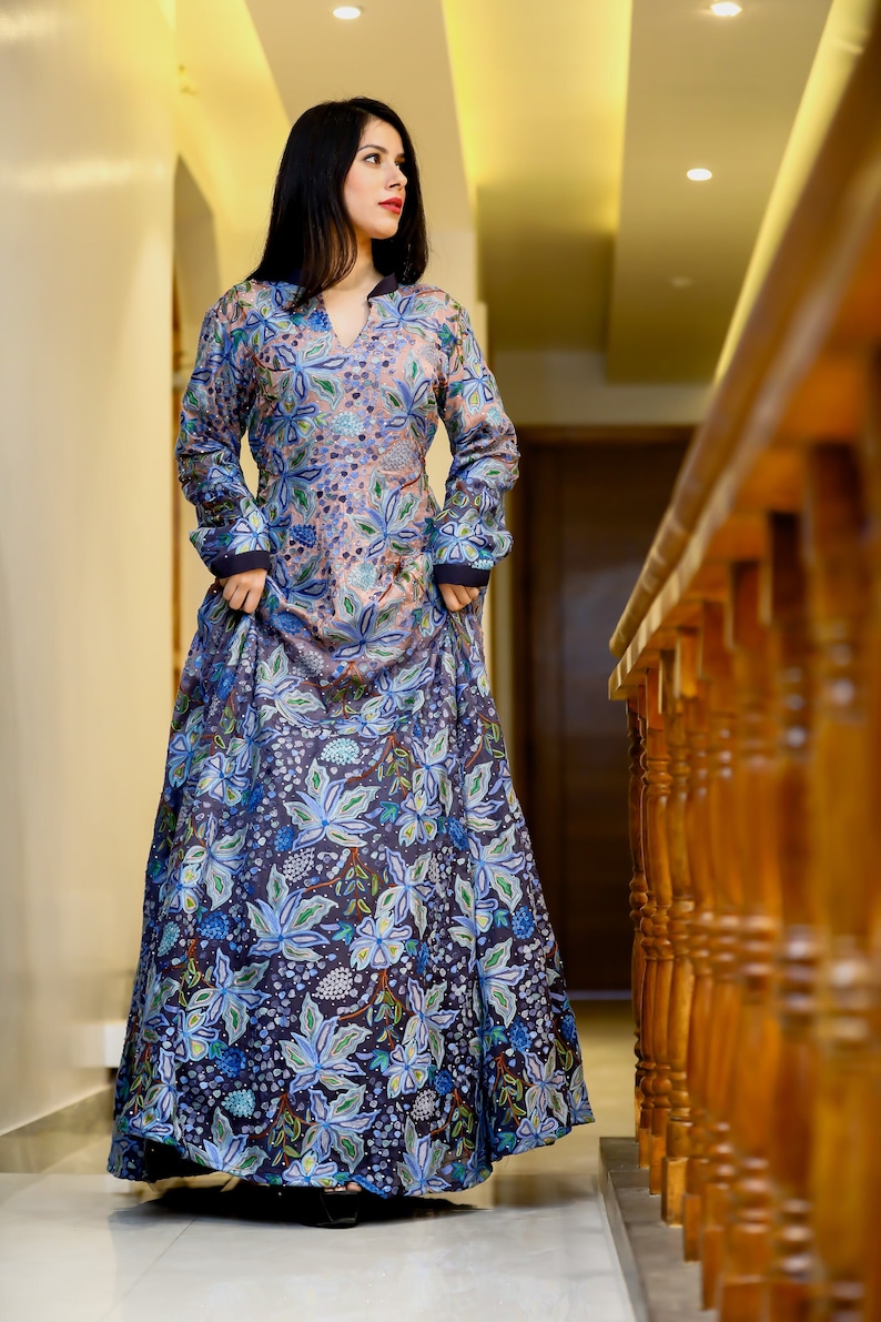 May include: A woman wearing a long, flowing floral dress with a blue and pink color scheme. The dress has a high neckline and long sleeves. The woman is standing in a hallway with a wooden railing in the background.