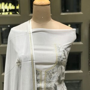 Op de afbeelding: Witte geborduurde jurk met zilveren draadborduurwerk. De jurk heeft lange mouwen en een hoge halslijn. De jurk is op een wit mannequin.