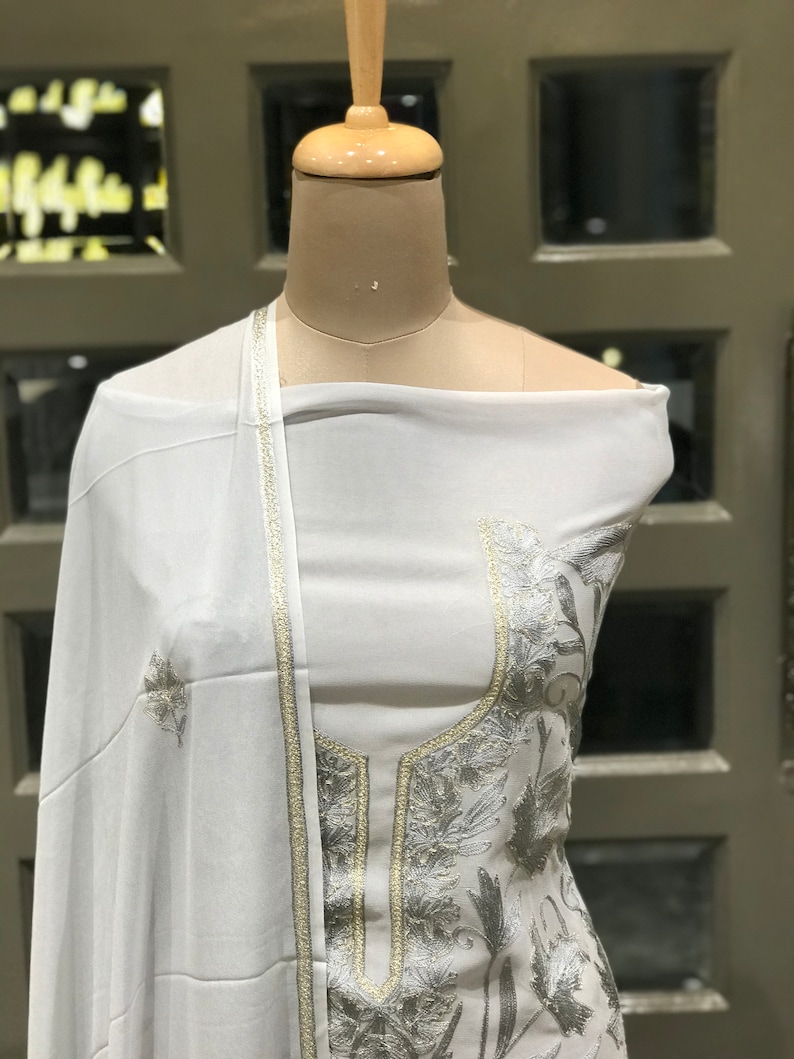 Op de afbeelding: Witte geborduurde jurk met zilveren draadborduurwerk. De jurk heeft lange mouwen en een hoge halslijn. De jurk is op een wit mannequin.