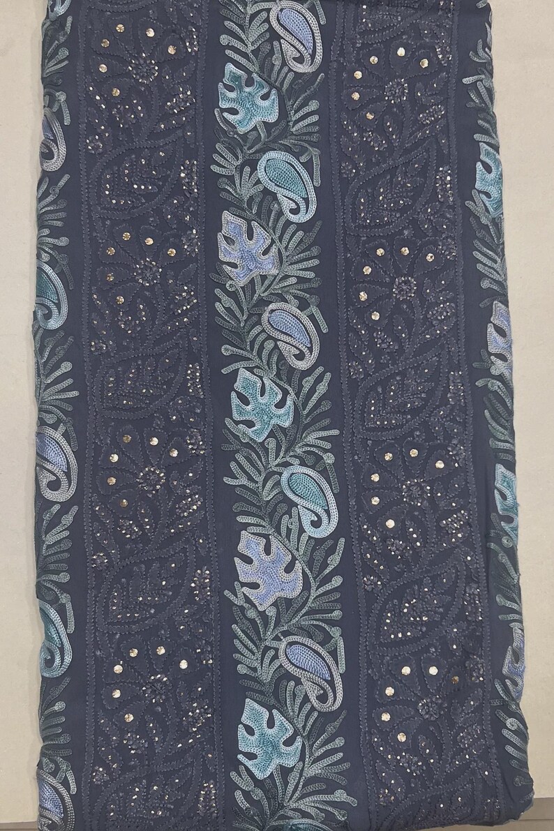 May include: A dark grey fabric featuring vertical panels of intricate embroidery. The design includes alternating bands of floral patterns with small metallic sequins and central columns decorated with light blue and teal leaf and paisley motifs.