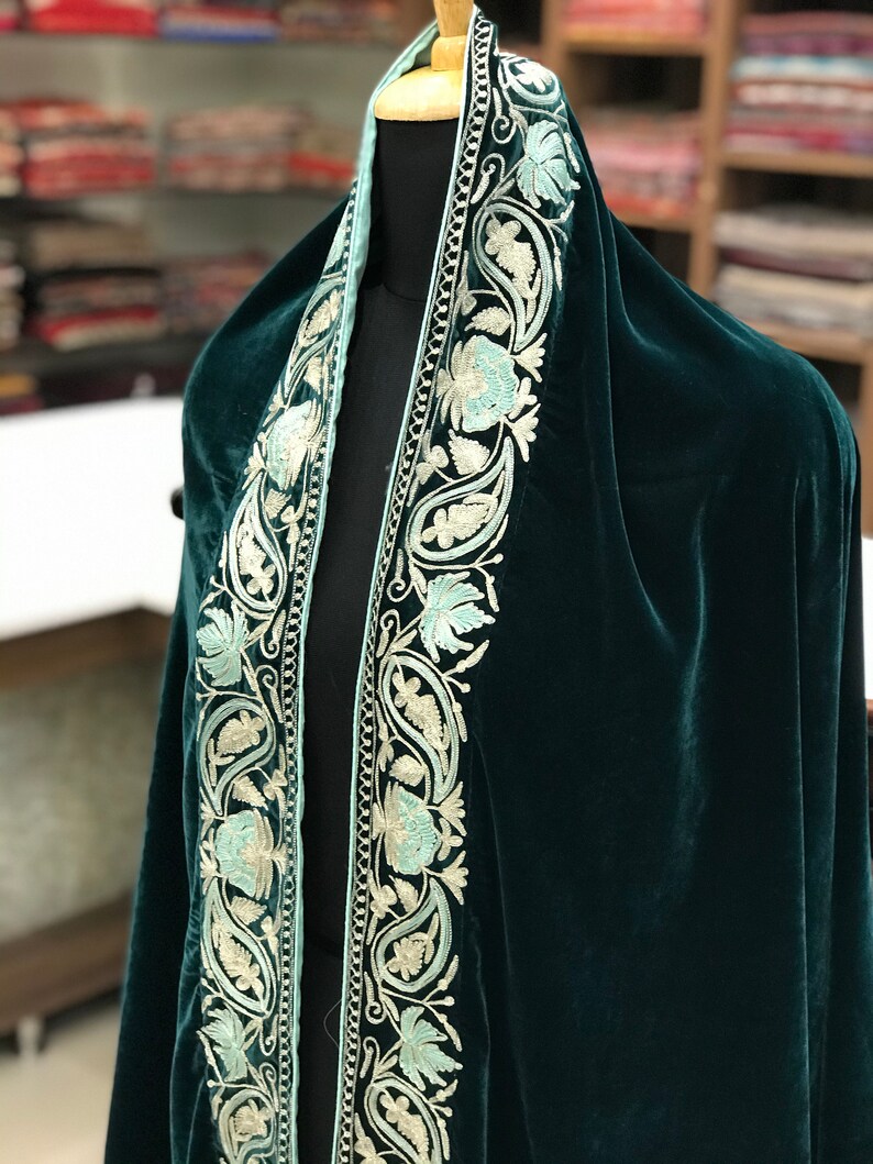 May include: A dark green velvet shawl with a light blue and white embroidered border. The embroidery features paisley and floral designs.