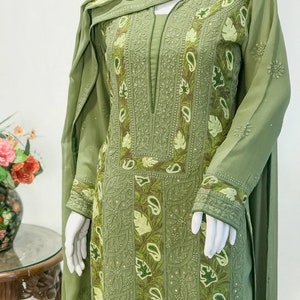 May include: An olive green long-sleeved tunic featuring intricate tonal embroidery, leaf motifs, and paisley patterns along the front panel and cuffs. The garment includes a matching olive green scarf draped over the shoulders, showcasing detailed needlework.