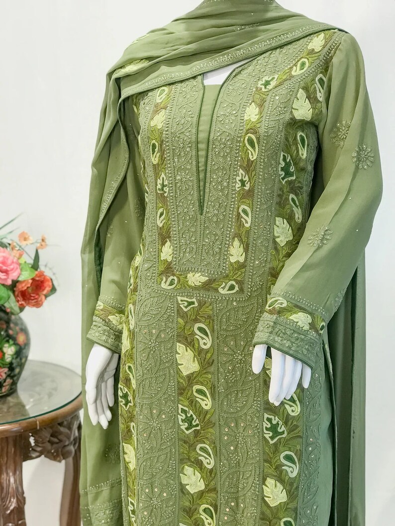 May include: An olive green long-sleeved tunic featuring intricate tonal embroidery, leaf motifs, and paisley patterns along the front panel and cuffs. The garment includes a matching olive green scarf draped over the shoulders, showcasing detailed needlework.