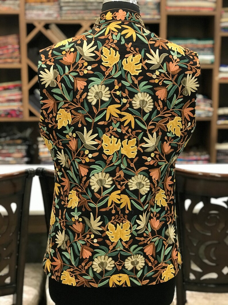 K&ouml;nnte beinhalten: Eine schwarze, &auml;rmellose Weste mit einem floralen Stickmuster in Gelb-, Gr&uuml;n-, Braun- und Rosat&ouml;nen.