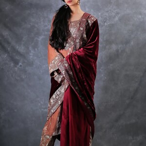 May include: A long-sleeved tunic and matching trousers set in a muted orange hue, featuring intricate silver embroidery on the bodice and pant cuffs. A deep burgundy velvet shawl is draped over one shoulder, complementing the ensemble's rich textures.
