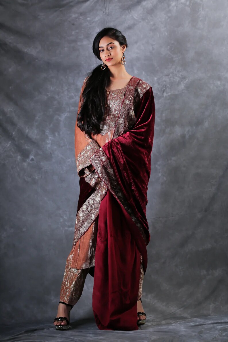 May include: A long-sleeved tunic and matching trousers set in a muted orange hue, featuring intricate silver embroidery on the bodice and pant cuffs. A deep burgundy velvet shawl is draped over one shoulder, complementing the ensemble's rich textures.