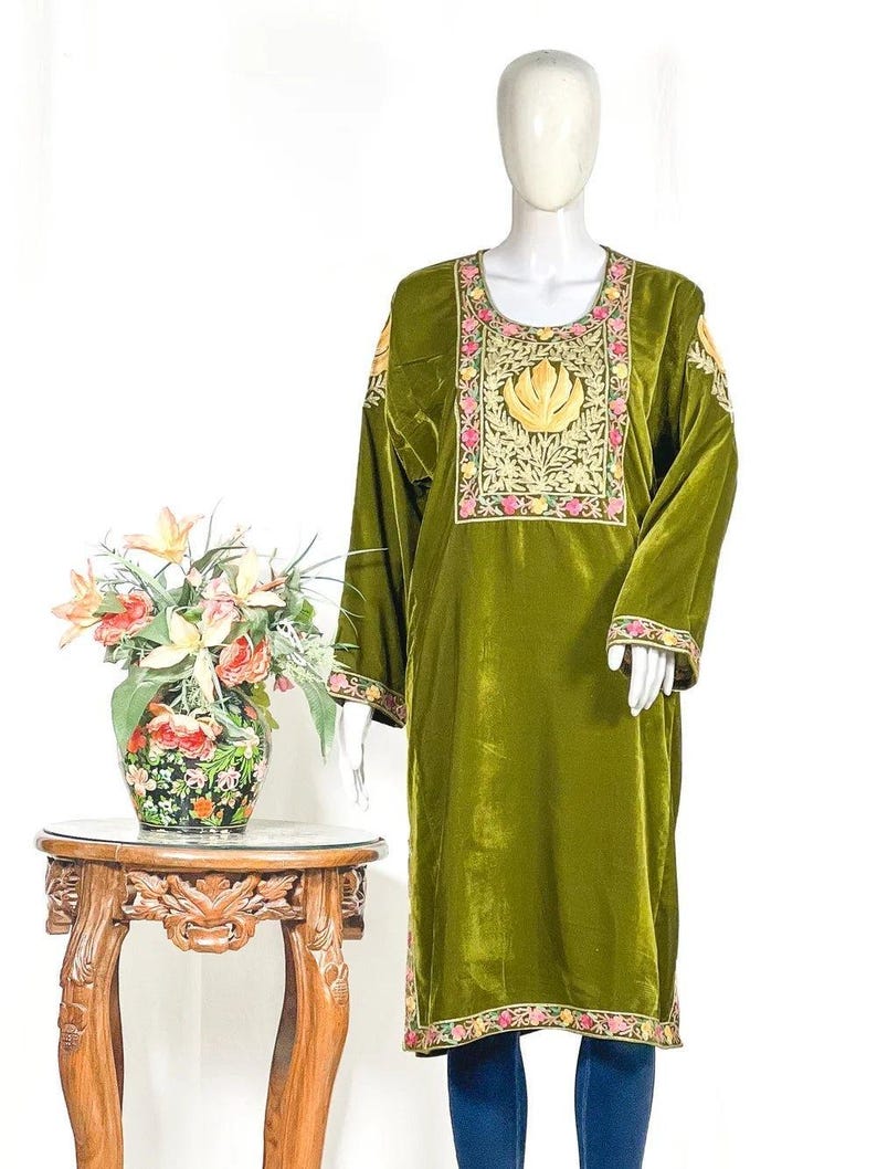 May include: An olive green velvet tunic featuring a scoop neckline and long sleeves. The garment is decorated with intricate floral embroidery in pink, yellow, and green thread around the neckline, cuffs, and hem, with a central golden leaf motif.
