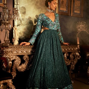 Pode incluir: Uma mulher vestindo um vestido azul-esverdeado com padr&atilde;o floral e decote profundo. O vestido tem mangas compridas e um corpete justo. A mulher est&aacute; em p&eacute; em um quarto com m&oacute;veis ornamentados e um espelho.