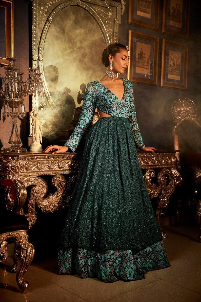 Pode incluir: Uma mulher vestindo um vestido azul-esverdeado com padr&atilde;o floral e decote profundo. O vestido tem mangas compridas e um corpete justo. A mulher est&aacute; em p&eacute; em um quarto com m&oacute;veis ornamentados e um espelho.