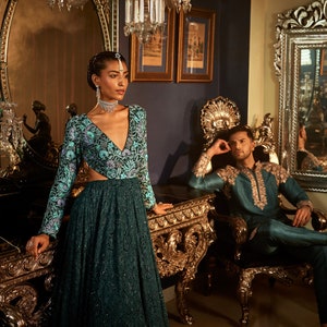Pode incluir: Uma mulher com um vestido estampado em azul e verde-azulado com um decote profundo est&aacute; em p&eacute; em frente a um espelho dourado adornado. Um homem com uma kurta azul-esverdeada com bordados dourados est&aacute; sentado em uma cadeira dourada adornada.