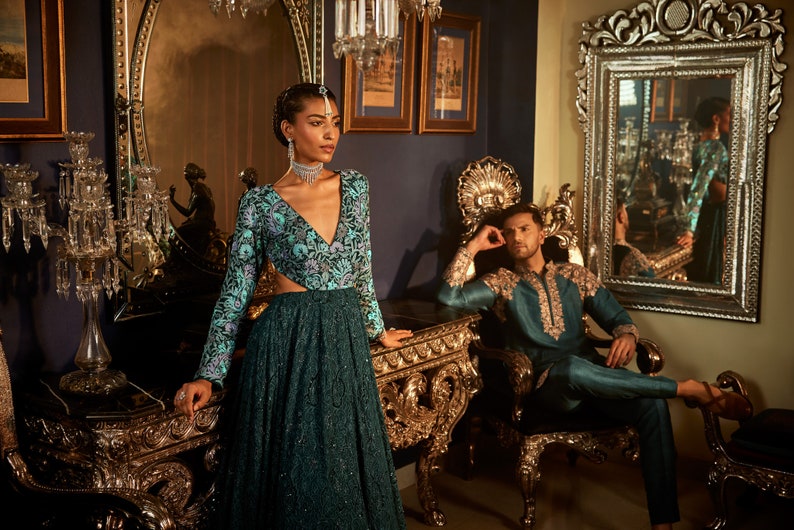 Pode incluir: Uma mulher com um vestido estampado em azul e verde-azulado com um decote profundo est&aacute; em p&eacute; em frente a um espelho dourado adornado. Um homem com uma kurta azul-esverdeada com bordados dourados est&aacute; sentado em uma cadeira dourada adornada.