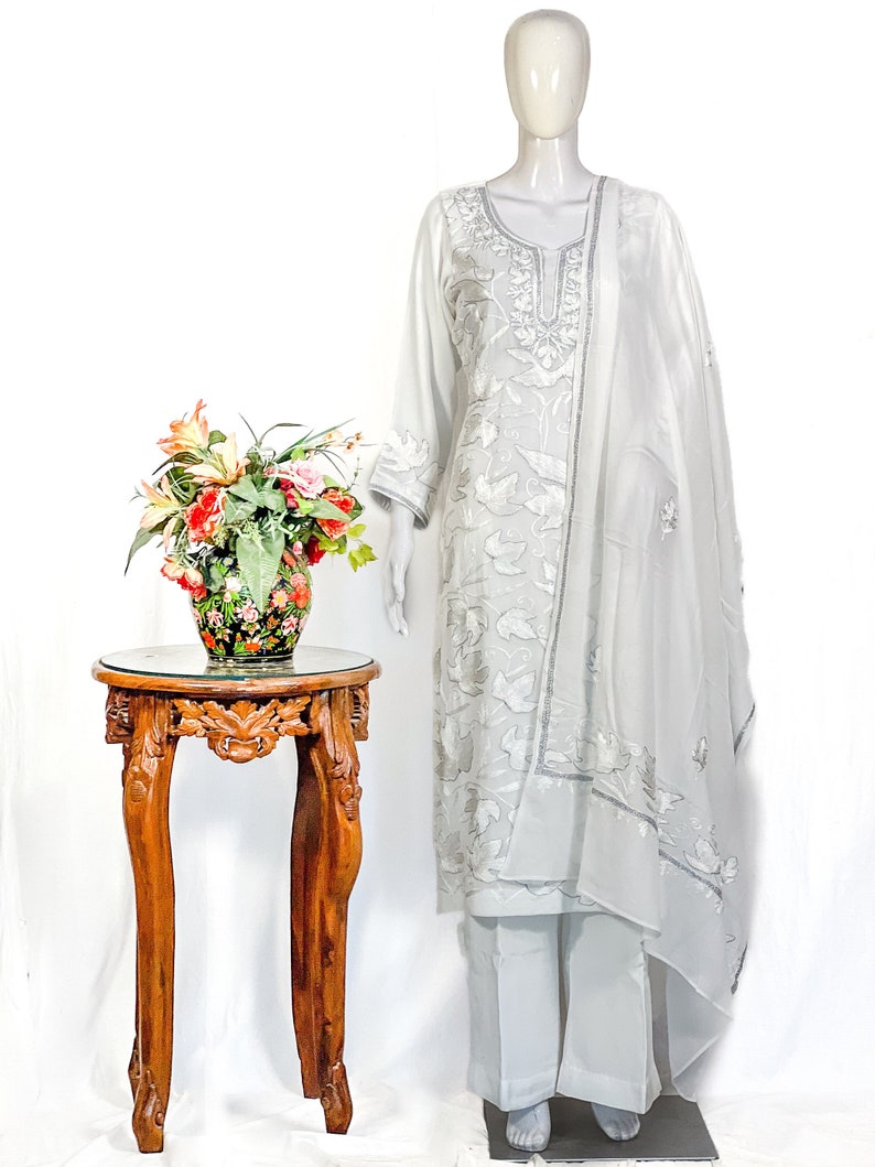 May include: A light gray three-piece outfit consisting of a long-sleeved tunic with silver floral embroidery, matching straight-leg trousers, and a coordinating sheer dupatta draped over the shoulder, all displayed on a mannequin.