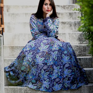 May include: A woman wearing a long, flowing blue dress with a floral pattern sits on a set of concrete stairs. The dress has a dark blue background with light blue, green, and white flowers.