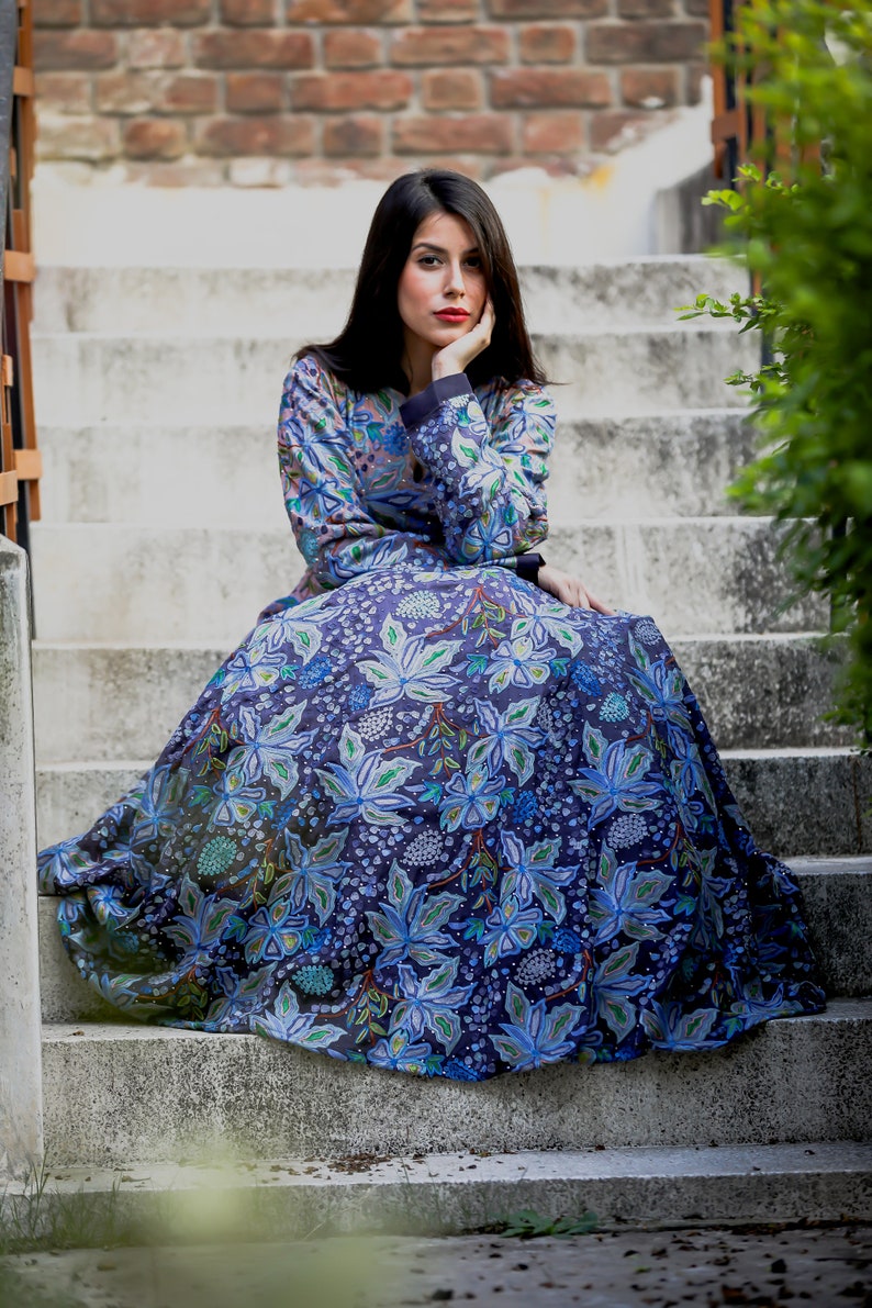 May include: A woman wearing a long, flowing blue dress with a floral pattern sits on a set of concrete stairs. The dress has a dark blue background with light blue, green, and white flowers.