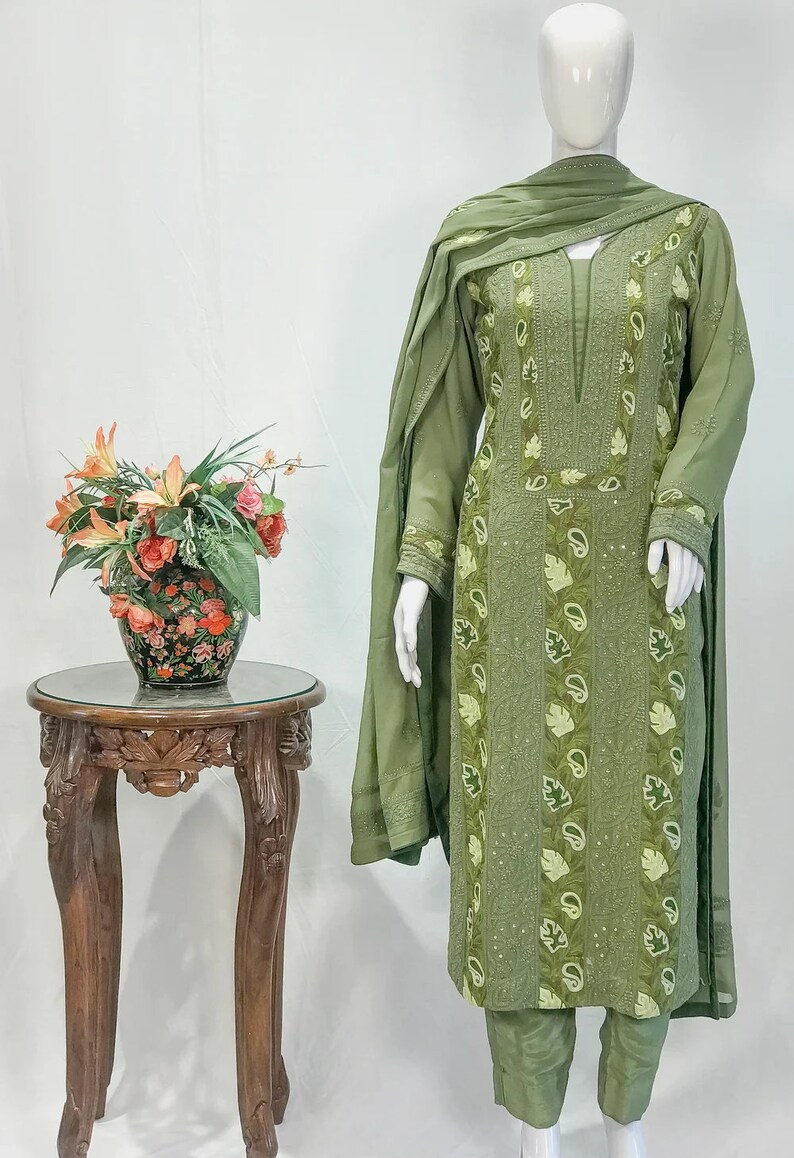 May include: An olive green three-piece outfit consisting of a long-sleeved tunic, trousers, and a matching dupatta. The tunic features intricate tonal embroidery, vertical panels with paisley and leaf motifs, and a notched neckline with subtle sequins.